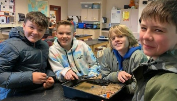 four boys smile for the camera at a lab table with a box of sand and materials